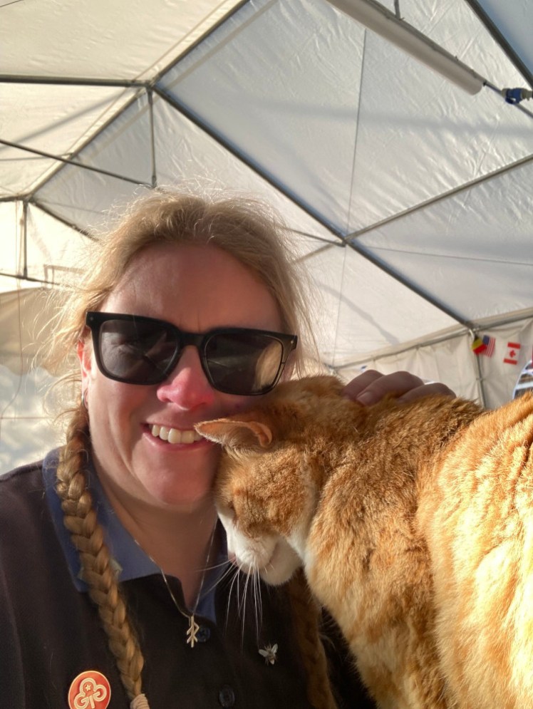 Me in my Guide uniform in a marquee wearing sunglasses at 7am. I'm sitting at a wooden picnic table and a ginger cat standing on the table is snuggling up against me.