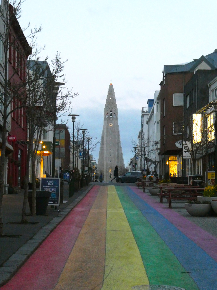 Looking up towards Hallgrimskirkja, a huge white space shuttle-shaped church at the top of a road painted with rainbow stripes.