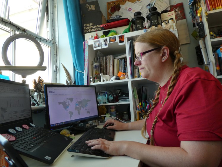 Me at my desk, wearing a red t-shirt and looking intensely at my laptop while a map of the world is visible on the second screen to my right. The desk is actually tidy but the Kallax unit behind me doesn't make it look that way.