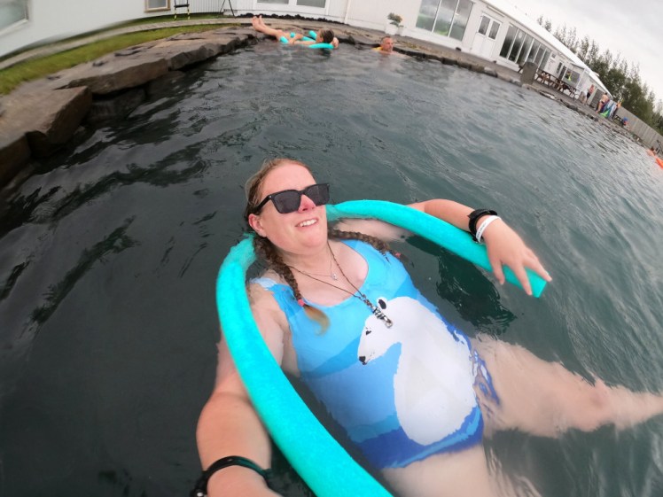 Selfie in the Secret Lagoon. I'm floating on my back with a foam noodle under my neck, wearing a blue striped swimsuit with a polar bear on it and also sunglasses.