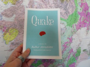 Quake by Auður Jónsdóttir, a book with a matte textured paper cover in mint green, with a tiny red brain in the middle and a wiggly line running through the title in white letters. I'm holding it up against my tectonic map of Iceland.