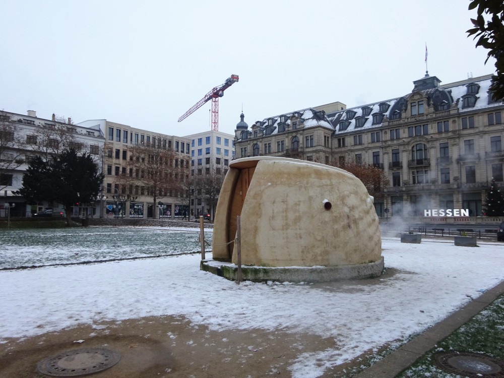 A large pebble-like building in the middle of a snowy square. Steam is billowing from the back of it and there are bare patches around steaming manholes.