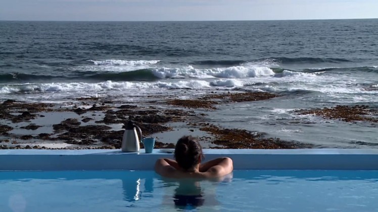 A woman at the end of a swimming pool, looking out across a seaweedy beach at the open ocean right by the pool. On the edge are a coffee pot and a mug.