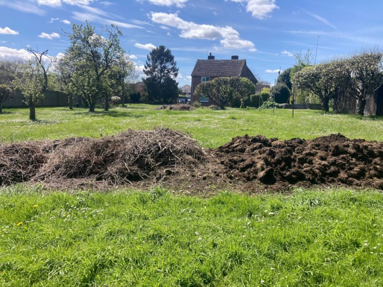 In the foreground, a vegetable patch is covered in manure. Behind it is a long garden and a grey house in the distance, behind some small trees.