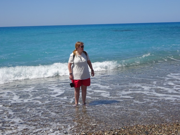 Me up to my ankles in the sea in Cyprus. I'm wearing a pale grey t-shirt and red shorts and have my camera dangling from my hand.