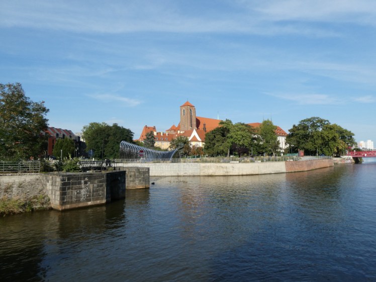 An island on the other side of the river with trees around the edge and church roofs and towers visible behind them.