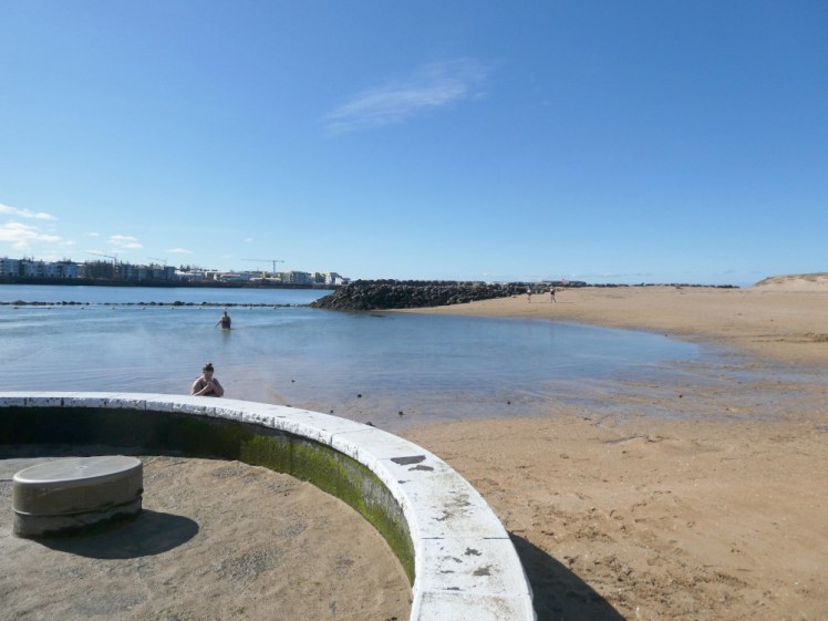 A sloping golden beach running down to a large pool of blue water, separated from the bay beyond by a rope. In the foreground is a round circular hotpot, currently just full of sand.