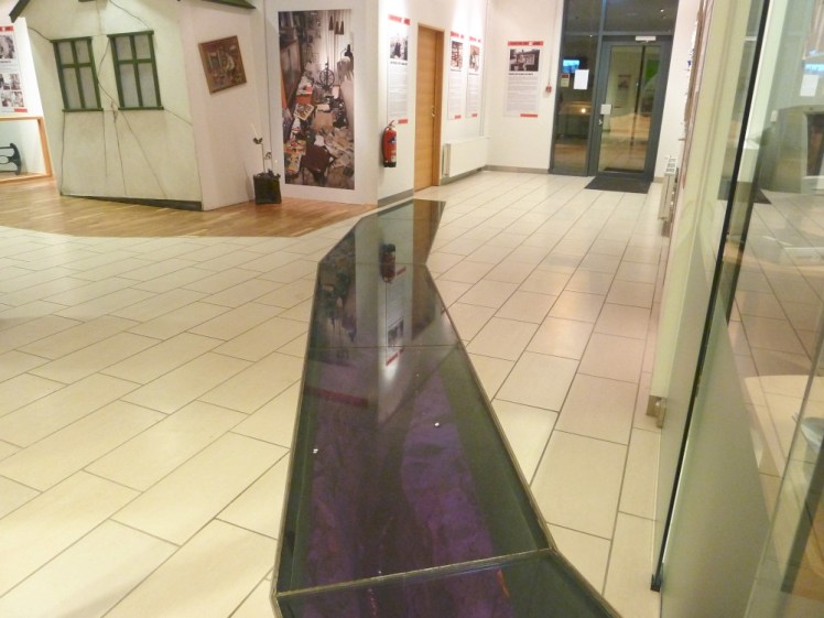 A shopping centre floor with a glass path in it, about two feet wide, following the route of a crack in the ground underneath. The crack is lit in purple.