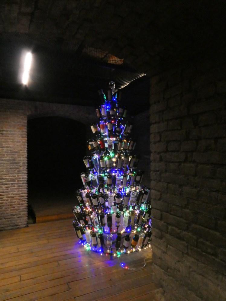 A Christmas tree made out of wine bottles and LEDs, in an underground wine museum with brick walls and ceiling and a fake wooden floor.