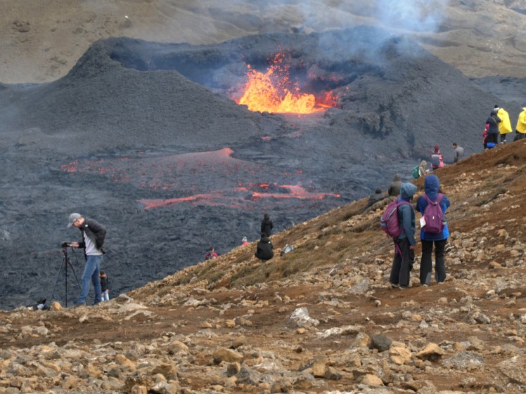 People in waterproof clothes and backpacks standing on a hillside. Behind them is a young fresh black volcano erupting orange and yellow lava. From this angle, it looks at least three times the size it actually is.