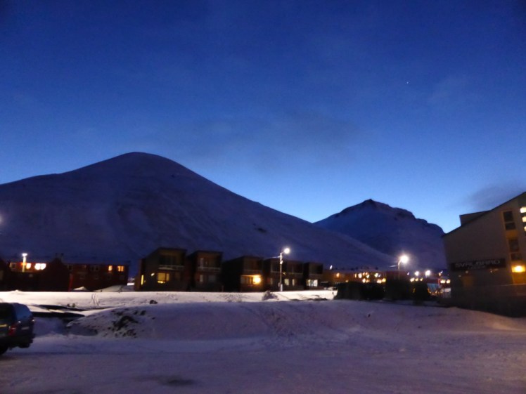 Svalbard in November. A small town sits at the foot of pointed mountains which are silhouetted by the dark blue sky. Anywhere else, this would be 30 to 60 minutes after sunset. This is about lunchtime in Svalbard in "dark winter".