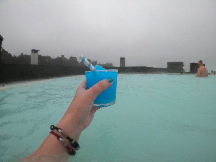 My hand holding a plastic glass of bright blue slush in the Blue Lagoon. The water is opaque greeny-blue and the sky is so cloudy you can't see anything.