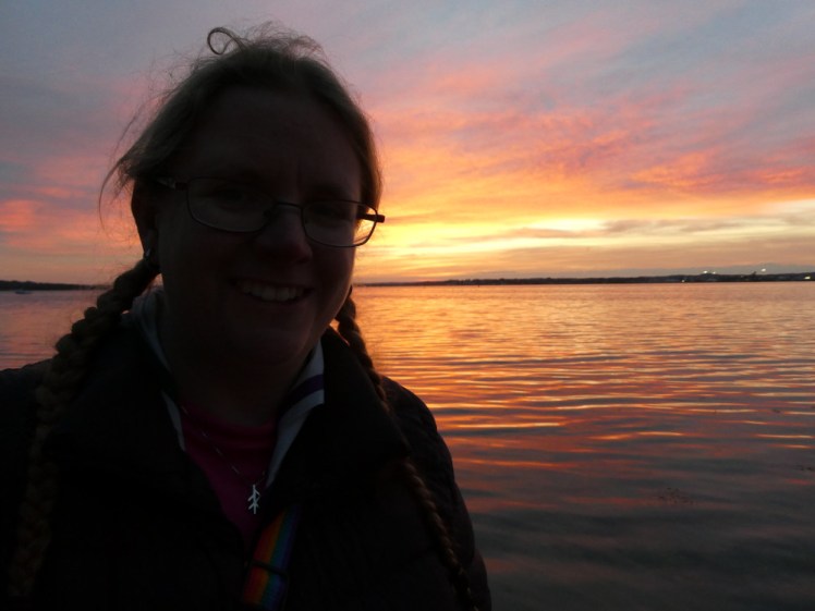 A selfie, almost silhouetted against a pink and orange sky which is reflecting pure orange off the sea behind me.