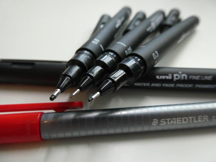 Four Uni Pin fineliners. Three with their lids off to show the differences in the fine nibs, balancing on the back of the fourth with a red Staed