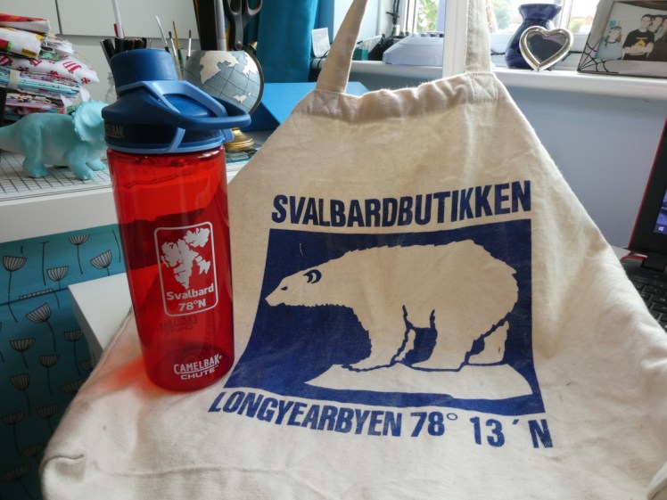 A red translucent water bottle with a blue lid and a white Svalbard island map and coordinate printed on the front, and a canvas shopping bag with the blue polar bear logo of Svalbardbutikken and coordinates.