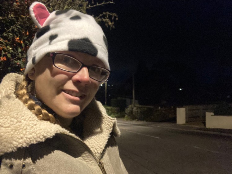 A selfie taken on my walk in the dark under a street light. I'm wearing a fleece cow-patterned hat complete with ears and am wearing a sheepskin poncho.