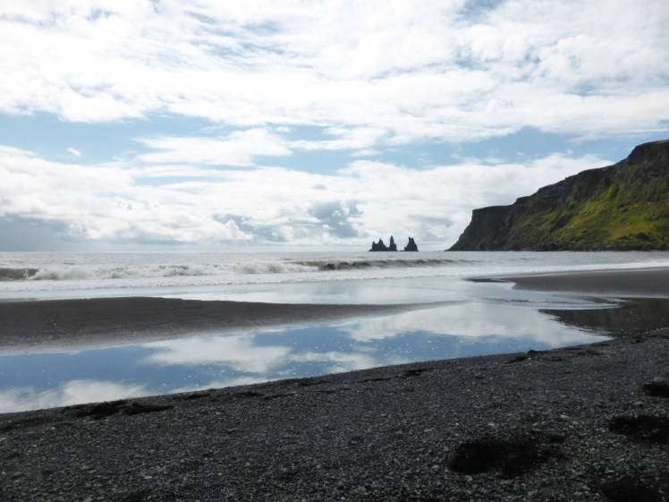 The black sand beach at Vik. In the foreground is black sand and between the sand and the rolling waves is a big patch of water reflecting the blue but cloudy sky. To the right are some mossy cliffs and at the end of them, some very spiky pinnacles out to sea.