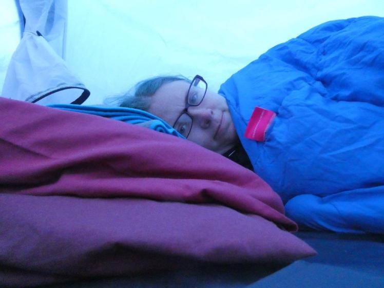 Selfie in my sleeping bag. Everything is blue-tinted by the blue tent fabric but the sleeping bag is blue and the pillows are reddish-purple.