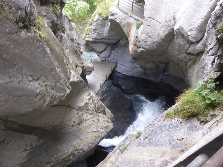 A waterfall from above. Most of the picture is of rock carved into rounded shapes by violent water over thousands of years. At the bottom, the water is seen gushing out from the left and running away into another carved tube somewhere.