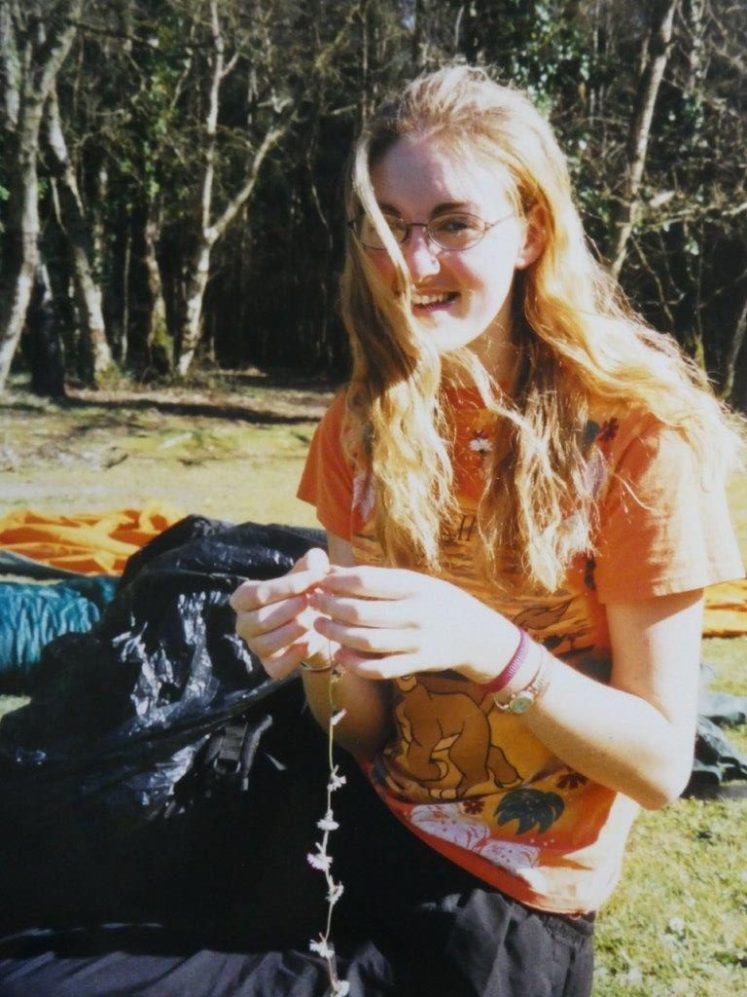 Vicky sitting on the grass making a daisy chain. Her hair is down too and she's laughing at the camera.