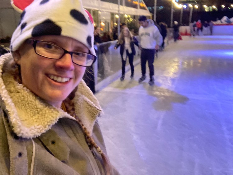 A selfie on the ice. You can't see my feet but you can see the ice directly behind me, and people staying very close to the wall. I'm wearing my cow-patterned hat and my fake sheepskin poncho and look pretty happy to be skating.