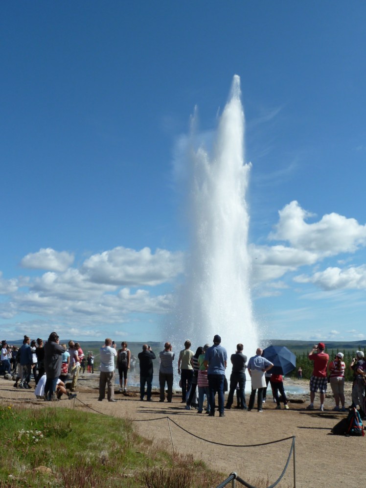 A portrait photo of a geyser erupting, with a ring of tourists standing in front of it.