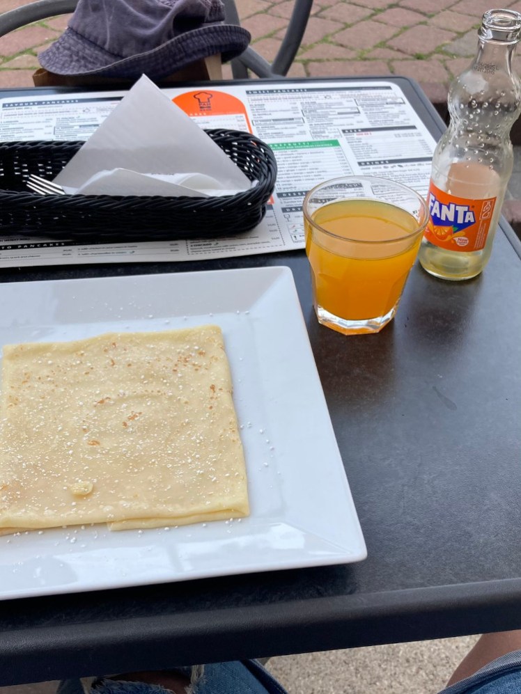 A square pancake with sugar on a square white plate. On the table is a small glass and a glass bottle of Fanta plus the menu behind it.