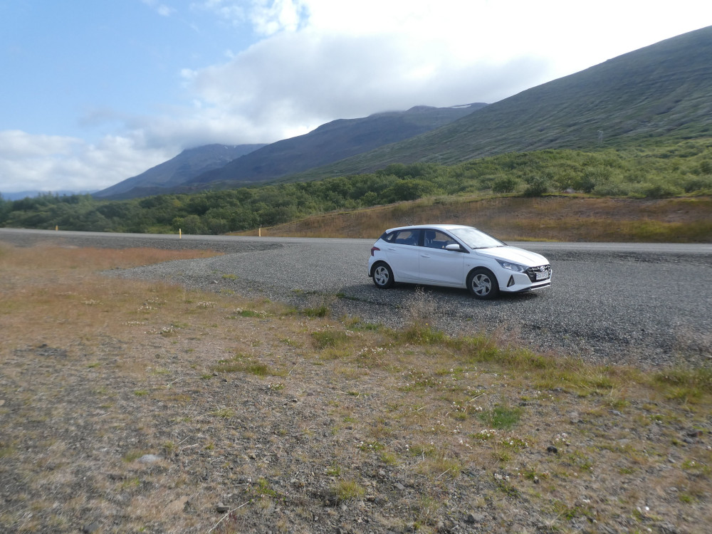 A white Hyundai i20 parked on the edge of a road through a gravelly yet grassy area of mountains.