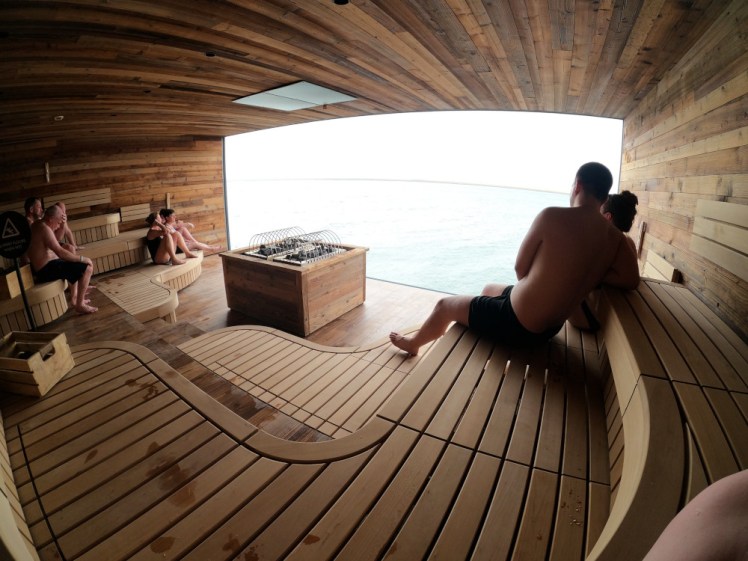 Inside the Sky Lagoon's sauna. A square room with pale wood walls and benches on three levels, a heating thing at the front and a huge perfect window.