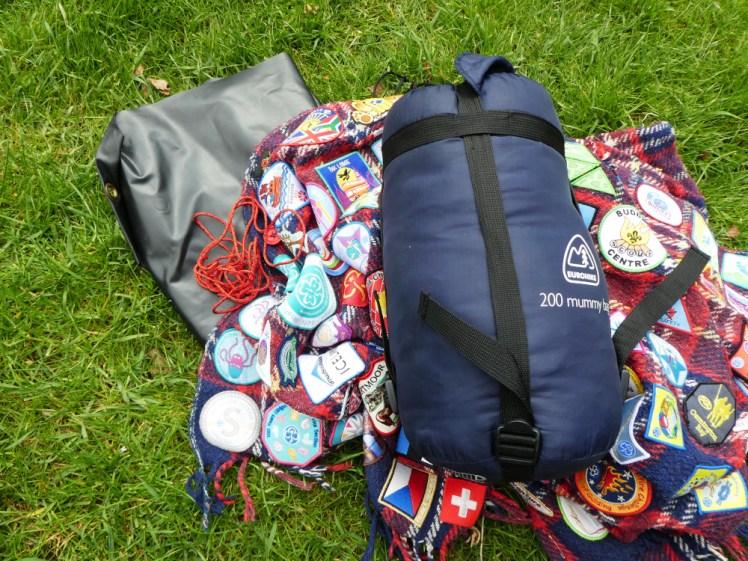 A pile of black groundsheet, red badge-covered blanket, sleeping bag in its stuffsack and handful of red cord lying in the grass.