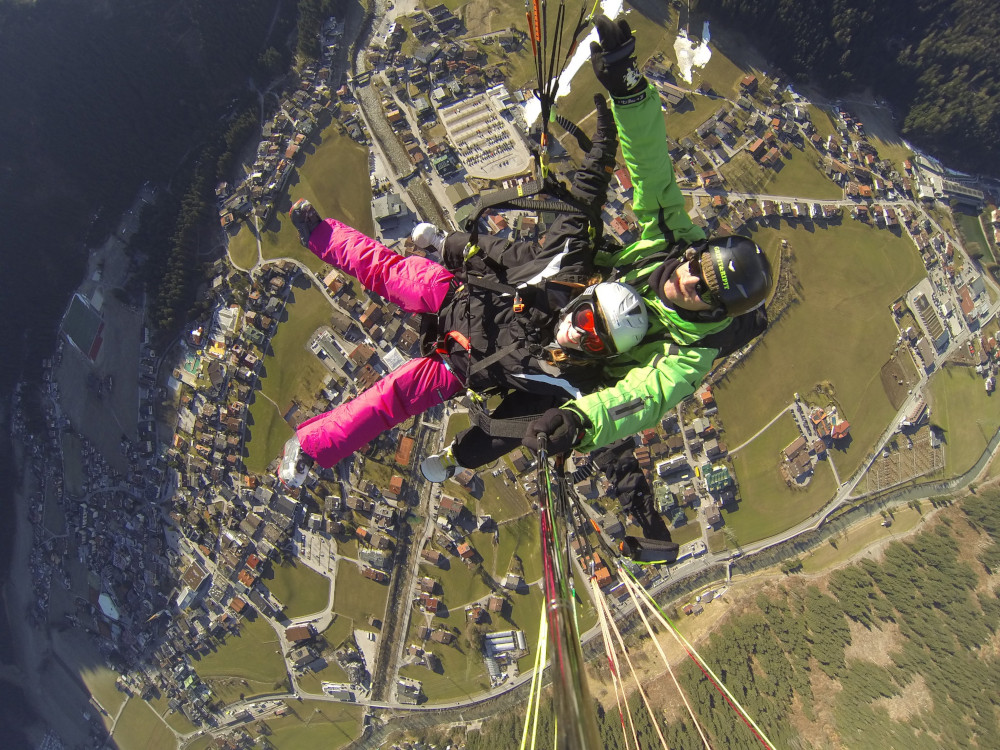A shot straight down from a selfie stick through the lines of a paragliding wing to me and my pilot. I'm wearing neon pink ski trousers and a black jacket, with arms and legs starfished out. He's wearing a neo green ski jacket and you can't see his legs because I'm pretty much sitting on them. Directly below us, give or take a couple of hundred (or maybe thousand) feet is the ski resort of Mayrhofen.