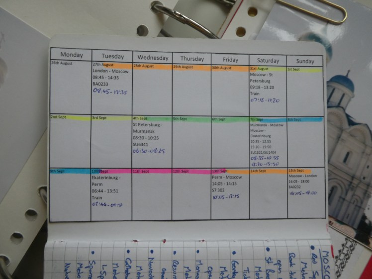 A calendar printed and stuck in the front of a small notebook. Days are colour-coded in highlighter depending on what city I'm in, and times and numbers show flights and trains. The times written in pen are conversions to UK time.