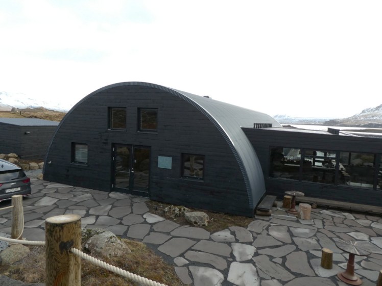 Hvammsvik building. A black round-roofed building like an oversized bunker, with a black-painted wooden front and a restaurant on the side. Out the front are irregular-shaped paving slabs and behind you can just make out the mountains around the fjord.