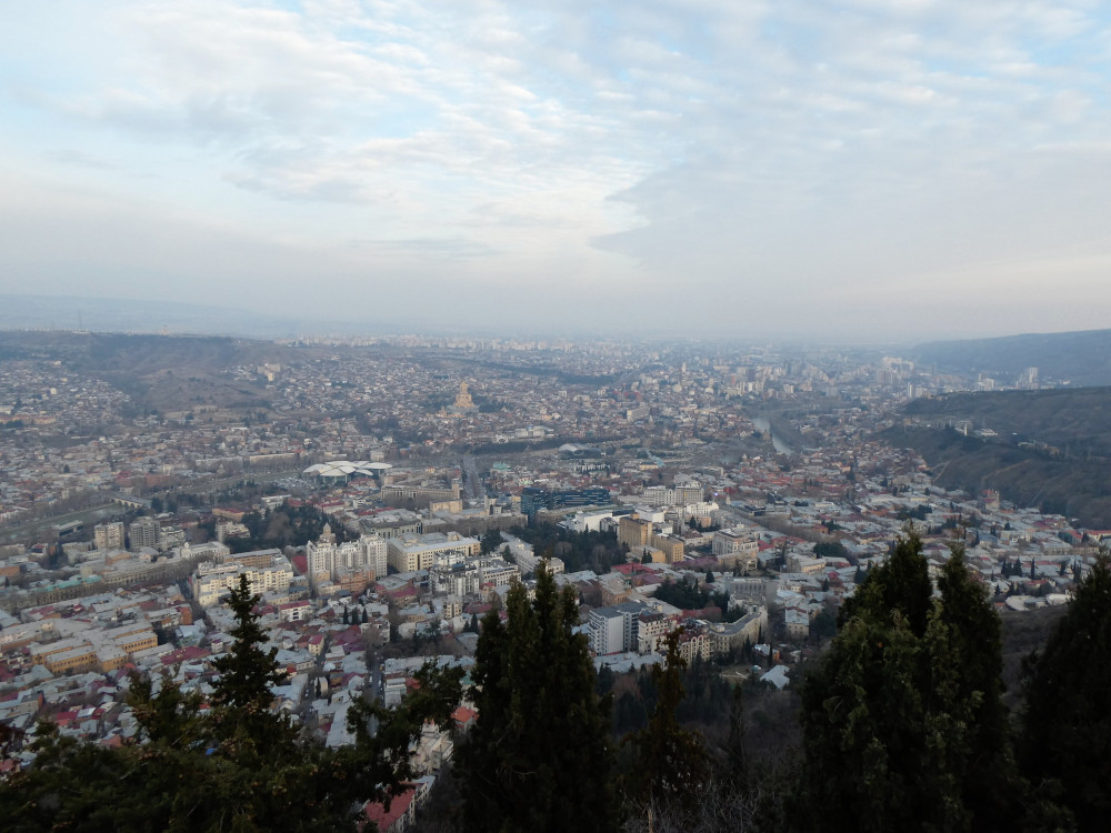 A slightly hazy evening view over Tbilisi from above, a sprawly city spreading out into the distance with the feet of mountains occasionally protruding into the urban sprawl.