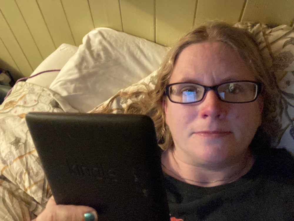 A selfie in bed holding up my Kindle. The reflection of whatever I'm reading is glowing blue in my glasses. My hair looks reasonably clean!