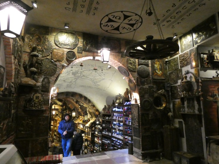 Inside the entrance of Meidan Bazar. An archway over stairs leads to the passageway and the walls are absolutely covered in brass plates, art etc. On the ceiling is the Georgian alphabet and a hanging cart wheel.