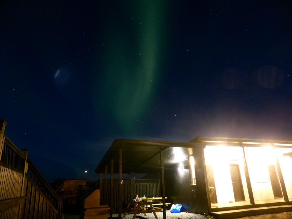 A long-exposure photo of a wooden campsite building (its lights aren't that bright, it's the long exposure) with a dark blue sky overhead. Streaking the middle of the picture is a bright band of green light, slightly more blue-tinted than the yellow-tinted greens you usually see.