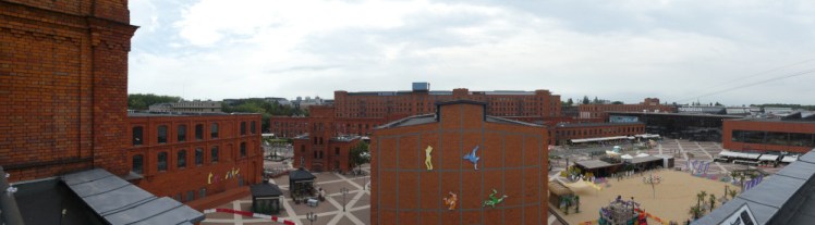 A panoramic view of the Manufaktura complex, showing red brick buildings of various shapes and sizes everywhere. To the right is the glass shopping centre built onto the red brick factory.