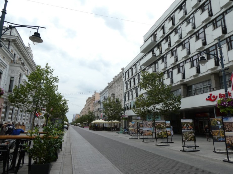A view down Piotrkowska. The most prominent building in this photo is a bank but there are small trees, pavement cafes and elsewhere, more decorative buildings. The road itself is absolutely flat grey brick with only a shallow gutter separating road from pedestrian walkway.