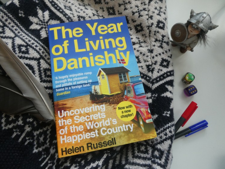 The Year of Living Danishly by Helen Russell - a book with a mostly blue and yellow cover lying on a black and white Nordic-style jumper, with a Scandinavian troll, the two colourful dice I bought in Denmark and a couple of pens sitting to the side.