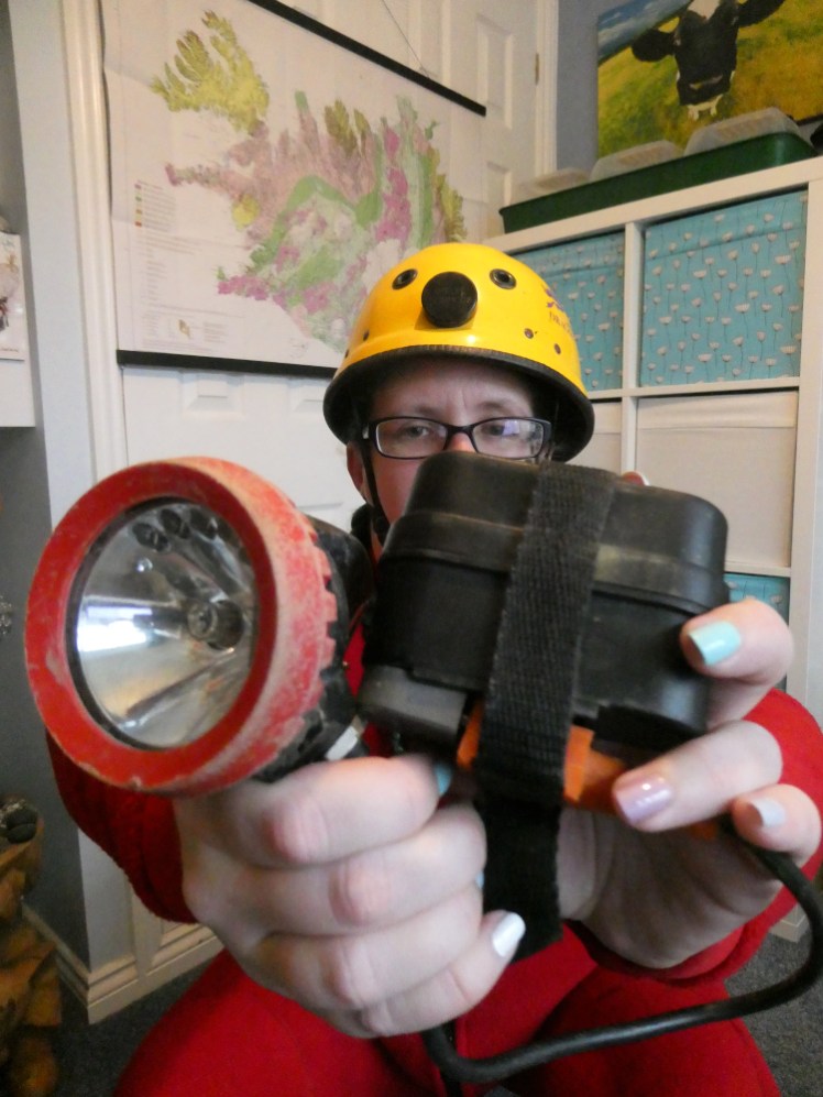 Me, in red undersuit and yellow helmet, holding my caving lamp and battery up to the camera. The lamp is a battered dusty piece with a red sealing ring and the battery is a rectangular black box with an orange plug joining the lamp to the battery.