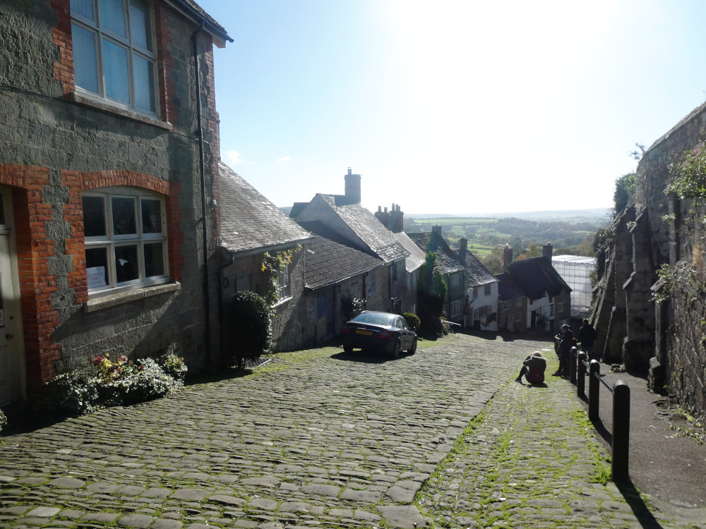 Gold Hill, a famous cobbled street with stone cottages down one side and a buttressed stone wall down the other. The sun is directly in front, washing out the sky and half-silhouetting the cottages.