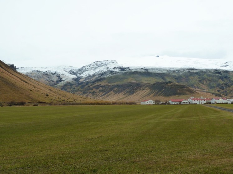 A grassy pasture leads to a farm composed of many buildings with white walls and red roofs. Immediately behind the farm is a mass of mountain capped with a mass of ice. This is where you'll have seen photos of the farm buried in ash from the 2010 eruption.
