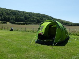My big green four-man tent pitched in a smooth green field. Behind is a hill covered in trees and the sky is blue.