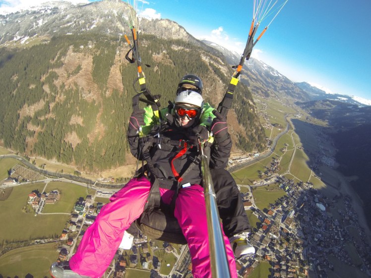 Me in bright pink ski trousers obscuring my pilot, hanging from a paraglider over Mayrhofen, a ski town in Austria. I don't know how high we are but we appear to be at least as high as the mountain behind the town.