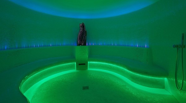The Alpine steam room, a white-tiled room lit in green and blue with a tiled bench around the edge of the walls.