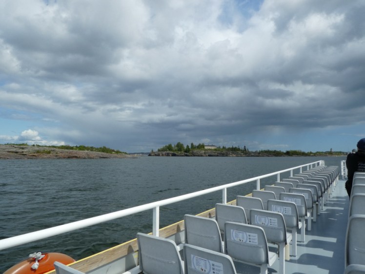 The upstairs of the boat, lots of seats in rows and a railing. In the background, there are rocky islands and a heavy cloud reflecting in the sea, making the water a dark grey-blue.