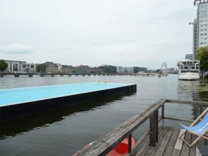 Badeschiff, a bright blue rectangular pool in the hull of an old barge, floating in a river. The river water is noticeably darker and slightly browner than the vivid turquoise pool water.
