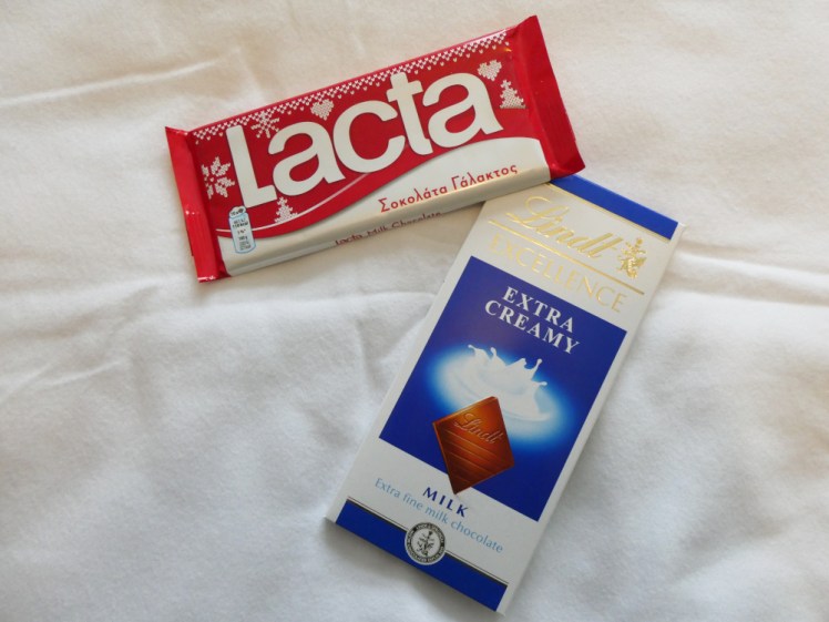 A bar of Greek Lacta chocolate, in a red plastic wrapper, next to Swiss Lindt Excellence, in a blue and white cardboard box.