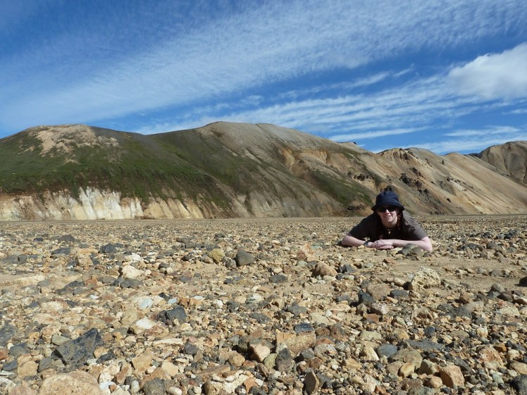Me lying in the gravel plain at Landmannalaugar, surrounded by yellow stripy grass-streaked mountains.